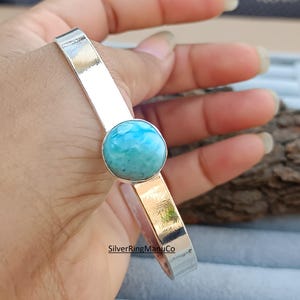 Könnte beinhalten: Ein silberfarbenes Armreif-Armband mit einem runden, hellblauen Edelstein. Der Edelstein ist in einer silbernen Lünette gefasst, und das Armband hat eine polierte Oberfläche. Das Armband ist ein Schmuckstück.