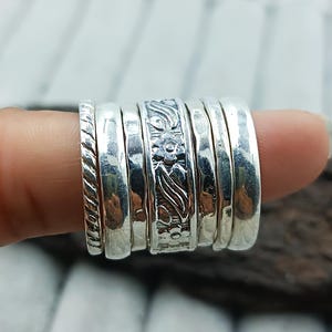 May include: A collection of silver rings, each with a unique design. The rings include a twisted rope pattern, hammered textures, and floral engravings. The rings are stacked together, showcasing the variety of styles and textures.