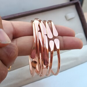 Könnte beinhalten: Eine Sammlung von roségoldenen Armreifen. Jeder Armreif hat ein tropfenförmiges Design an der Oberseite mit glatter, polierter Oberfläche. Die Armreifen sind gestapelt und zeigen ihre reflektierenden Oberflächen und ihren eleganten Stil.