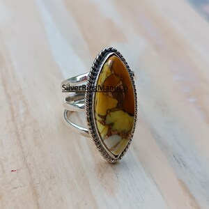 Könnte beinhalten: Ein silberner Ring mit einem ovalen Stein. Der Stein hat eine Mischung aus Gelb, Braun und Weiß. Der Ring hat einen geflochtenen Rand und mehrere Bänder. Der Text "SilverRingManuCo" ist auf dem Ring sichtbar.