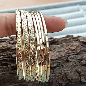 Op de afbeelding: Een verzameling gouden armbanden, sommige met bloemmotieven, wordt getoond. De armbanden zijn op een stuk hout gestapeld. De armbanden hebben een glanzende goudkleur.