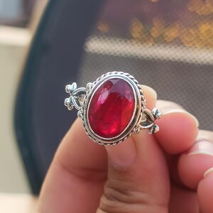 Könnte beinhalten: Ein silberner Ring mit einem ovalen roten Edelstein, der in einer dekorativen silbernen Fassung sitzt. Der Ring hat aufwendige florale Details an den Seiten.