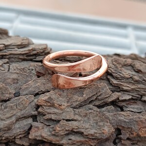 Könnte beinhalten: Ein kupferfarbener Ring mit einem einzigartigen, offenen Design. Der Ring umschließt den Finger mit einer glatten, polierten Oberfläche. Der Ring wird auf einer strukturierten, braunen Rindenoberfläche präsentiert.