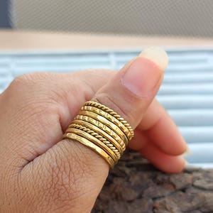 Könnte beinhalten: Ein Stapel goldfarbener Ringe an einem Daumen. Die Ringe haben verschiedene Texturen, darunter glatte, seilartige und geätzte Designs. Die Ringe sind übereinander gestapelt, wodurch ein geschichteter Effekt entsteht. Der Hintergrund ist verschwommen, mit einem Hauch von Holz.