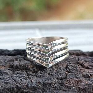 Peut inclure: Une bague en argent avec un motif en chevrons. La bague présente plusieurs couches, chacune avec un motif en forme de V. La bague est posée sur une surface sombre et texturée, avec un arrière-plan flou.