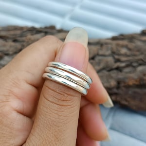 Könnte beinhalten: Ein Stapel von drei silbernen Ringen mit gehämmerten Texturen. Die Ringe werden auf einem Finger präsentiert und zeigen ihr schlichtes, aber elegantes Design. Die Ringe bestehen aus glänzendem, reflektierendem Metall.