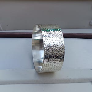 Könnte beinhalten: Ein silbernes Armreif-Armband mit einer strukturierten Oberfläche. Das Armband ist aus gehämmertem Metall mit einem kreisförmigen Muster.