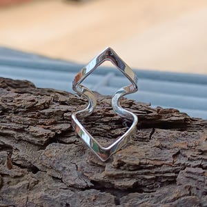Op de afbeelding: Een zilveren ring met een uniek ontwerp. De ring heeft twee ruitvormige elementen die zijn verbonden door een golvende, gebogen lijn. De ring is gemaakt van glanzend metaal en ligt op een stuk getextureerd hout.