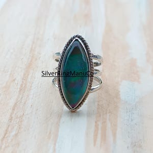Könnte beinhalten: Silberring mit einem grün schillernden Stein in Marquiseform. Der Stein ist von einem gedrehten Seildesign umgeben und auf einem mehrfach gebogenen Ring angebracht. Der Text "SilverRingManuCo" ist sichtbar.