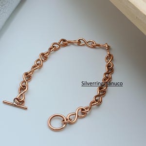 Könnte beinhalten: Ein kupferfarbenes Armband mit einem Kettendesign mit Unendlichkeitszeichen. Das Armband hat einen T-Bar-Verschluss und einen kreisförmigen Ringverschluss. Der Schmuck wird auf einer weißen Oberfläche präsentiert. Der Text "Silverringmanuco" ist sichtbar.