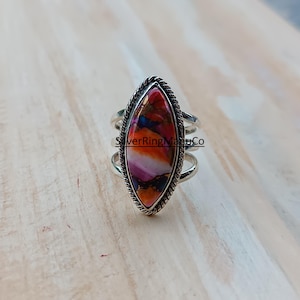 Könnte beinhalten: Ein silberner Ring mit einem ovalen Stein. Der Stein zeigt eine lebendige Mischung aus Farben: Rot, Orange, Lila und Weiß. Der Ring hat einen geflochtenen Rand und ein dreifaches Banddesign. Schmuck.