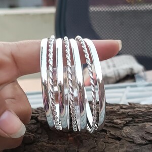 Puede incluir: Un conjunto de brazaletes plateados, algunos lisos y otros con un diseño de cuerda trenzada. Los brazaletes se exhiben sobre un trozo de madera, mostrando su superficie reflectante.