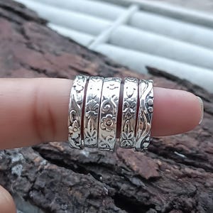 Könnte beinhalten: Fünf silberne Ringe mit Blumen- und Blättermustern werden auf einem Finger gezeigt. Die Ringe haben ein detailliertes, graviertes Design. Die Ringe bestehen aus einem glänzenden, metallischen Material. Der Hintergrund ist eine verschwommene Holzoberfläche.