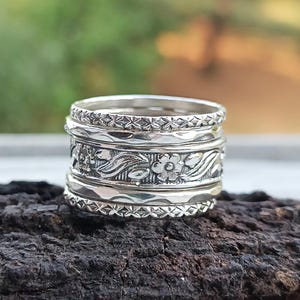 May include: A stack of silver rings with intricate floral and geometric patterns. The rings are stacked together, showcasing detailed designs of flowers, leaves, and repeating shapes. The rings are made of a shiny, reflective metal.