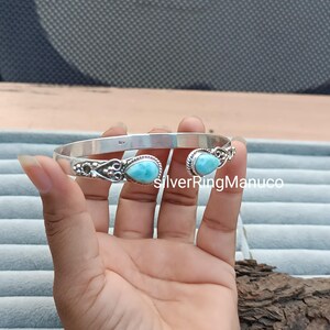 Op de afbeelding: Een zilveren manchetarmband met twee ovale, lichtblauwe edelstenen. De armband heeft een open ontwerp met decoratieve filigraan details bij de edelstenen. De tekst "SilverRingManuco" is zichtbaar.