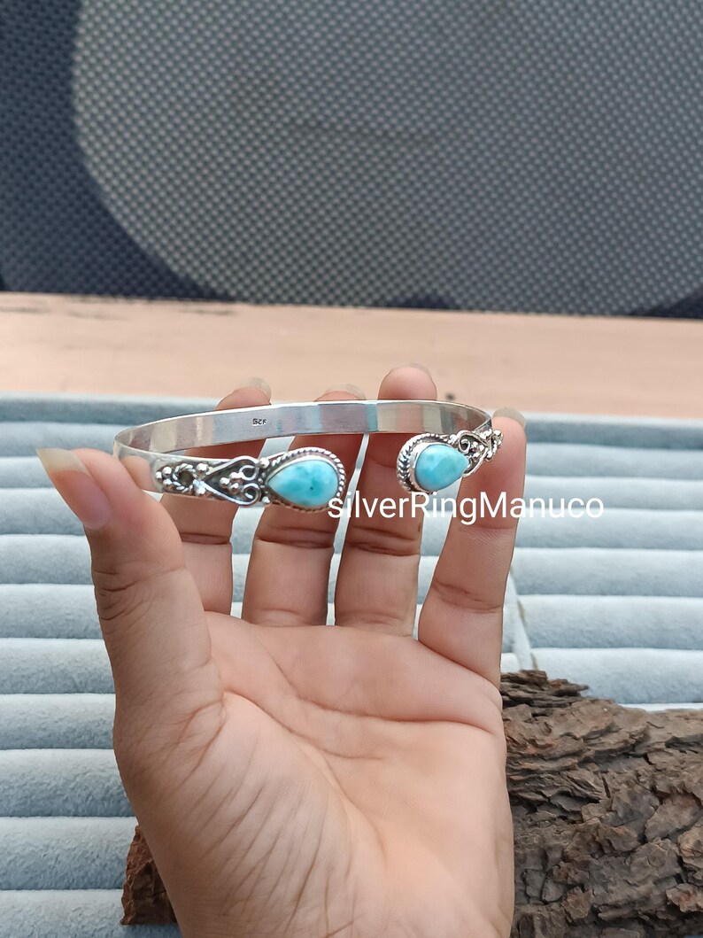 Op de afbeelding: Een zilveren manchetarmband met twee ovale, lichtblauwe edelstenen. De armband heeft een open ontwerp met decoratieve filigraan details bij de edelstenen. De tekst "SilverRingManuco" is zichtbaar.