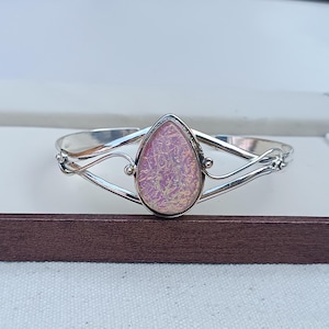 Op de afbeelding: Zilverkleurige manchetarmband met een peervormige, iriserende roze edelsteen. De armband heeft een delicaat, open ontwerp met wervelende details en kleine decoratieve accenten. Een stijlvol sieraad.
