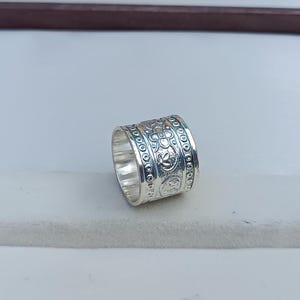 Könnte beinhalten: Ein silberner Ring mit einem breiten Band, das mit einem komplizierten, geprägten Design aus Kreisen und Blumenmustern verziert ist.