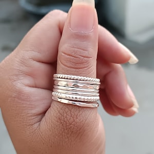 Puede incluir: Un conjunto de anillos de plata de diferentes diseños apilados en un dedo. Los anillos presentan diseños como cuerda trenzada, texturas martilladas y bandas lisas. Los anillos se muestran en un dedo, destacando la variedad de texturas y estilos.