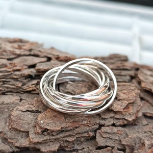 Op de afbeelding: Een zilveren ring met meerdere in elkaar grijpende banden. De ring is gemaakt van glanzend metaal en ligt op een getextureerd, bruin oppervlak. De banden zijn met elkaar verweven, wat een uniek ontwerp creëert. De ring is een sieraad.