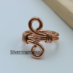 Puede incluir: Un anillo de alambre de color cobre con un diseño único y arremolinado. El anillo presenta una doble banda y un diseño abierto ajustable. El texto "Silverringmanuco" está impreso en el anillo.