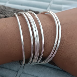 May include: A set of silver bangles. The bangles are thin, round, and shiny, reflecting light. The bracelets are stacked on top of each other, creating a layered effect. The jewelry is displayed on a person's arm.