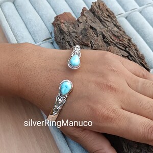 Op de afbeelding: Een zilveren manchetarmband en ring set, elk met een lichtblauwe, druppelvormige edelsteen. De armband en ring hebben decoratieve zilveren details. De open manchetarmband is verstelbaar. De ring heeft een bijpassend ontwerp.