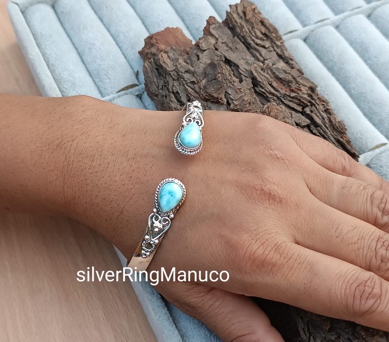 Op de afbeelding: Een zilveren manchetarmband en ring set, elk met een lichtblauwe, druppelvormige edelsteen. De armband en ring hebben decoratieve zilveren details. De open manchetarmband is verstelbaar. De ring heeft een bijpassend ontwerp.