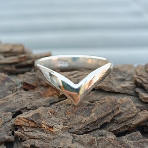 Könnte beinhalten: Ein silberner Ring in V-Form. Der Ring hat eine polierte Oberfläche und ein glattes, abgerundetes Band. Die V-Form ist zentriert und zeigt nach unten. Der Ring wird auf einer strukturierten, braunen Oberfläche präsentiert.