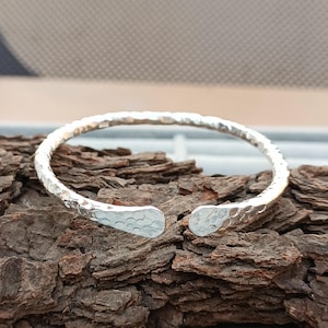 Könnte beinhalten: Ein silberfarbenes Armreif-Armband mit gehämmertem Muster. Das offene Armband hat eine strukturierte Oberfläche und abgerundete Enden. Das Armband wird auf einem Stück dunkelbraunem Holz präsentiert.