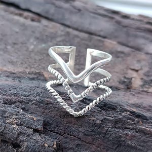 Puede incluir: Un anillo de plata con un diseño en capas. El anillo presenta tres bandas en forma de cheurón, con una textura trenzada similar a una cuerda en la banda inferior. La banda superior tiene un diseño recortado. El anillo se muestra sobre una superficie oscura y texturizada.