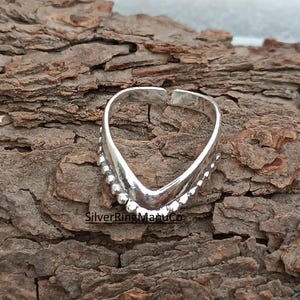 Peut inclure: Une bague argentée avec un design en forme de V. La bague présente une série de petites perles rondes le long du V. Elle est posée sur un fond en bois brun texturé. Le texte "SilverRingManuCo" est visible.