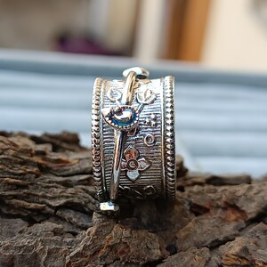 Könnte beinhalten: Ein silberfarbener Ring mit aufwendigen floralen und geometrischen Mustern. Der Ring hat ein drehbares Band mit detailliertem Design, einschließlich kleiner Blumen und Punkten. Der Ring ist auf einer strukturierten, dunkelbraunen Oberfläche platziert.