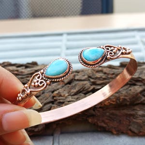 Könnte beinhalten: Kupfer-Armreif mit zwei türkisblauen, tropfenförmigen Steinen. Die Steine sind in aufwendigen, detaillierten Kupferfassungen gefasst. Das Armband hat ein offenes Design und wird von einer Hand gehalten.
