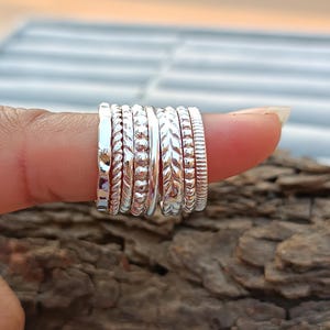 Könnte beinhalten: Ein Satz silberner Stapelringe mit verschiedenen Texturen und Mustern. Die Ringe sind auf einem Finger gestapelt und aus glänzendem Metall gefertigt.