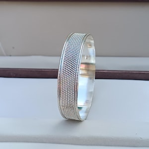 Könnte beinhalten: Ein silbernes Armreif-Armband mit einem strukturierten Design. Das Armband ist aus poliertem Silber gefertigt und hat eine glatte, abgerundete Kante.