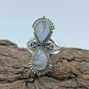Könnte beinhalten: Ein Silberring mit zwei tropfenförmigen Mondstein-Cabochons. Die Steine sind in verzierten, strukturierten Fassungen gefasst. Der Ring hat ein Doppelband-Design und wird auf einem Holzstück präsentiert.