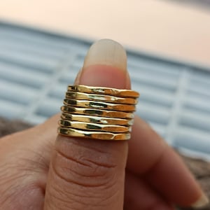 Puede incluir: Pila de anillos dorados. Los anillos son finos y tienen una textura martillada. Los anillos están apilados en un dedo. Los anillos son brillantes y reflejan la luz.