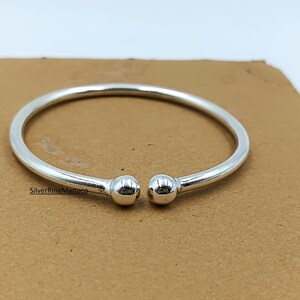 Könnte beinhalten: Ein poliertes, silberfarbenes Armband in offener Form mit einem glatten, kreisförmigen Band, das in zwei kleinen, abgerundeten, kugelförmigen Enden endet. Die Metalloberfläche ist reflektierend und einheitlich in der Farbe.