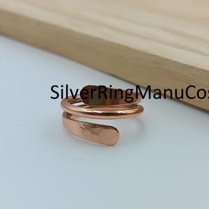 Könnte beinhalten: Ein kupferfarbener Bypass-Ring mit glatter, polierter Oberfläche und offenem, verstellbarem Design. Der Ring hat eine Spiralform mit zwei überlappenden Enden. Der Text SilverRingManuCos ist in schwarzer Schrift über dem Ring zu sehen.