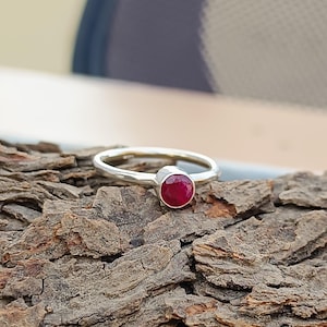 Könnte beinhalten: Ein silberner Ring mit einem runden, tiefroten Edelstein. Der Ring liegt auf einer strukturierten, braunen Oberfläche. Der Edelstein ist der Blickfang, mit einer polierten, reflektierenden Oberfläche. Das Band ist dünn und schlicht und hebt die Farbe und Form des Steins hervor.