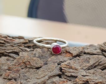 May include: A silver ring with a round, deep red gemstone. The ring is set on a textured, brown surface. The gemstone is the focal point, with a polished, reflective surface. The band is thin and simple, highlighting the stone's color and shape.