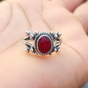 Könnte beinhalten: Ein silberner Ring mit einem roten Edelstein, der in einer dekorativen ovalen Fassung sitzt. Der Ring hat ein detailliertes Design mit kleinen Perlen um die Fassung.
