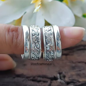 Könnte beinhalten: Nahaufnahme von vier Silberringen. Zwei Ringe haben ein Blumendesign vor dunklem Hintergrund. Die anderen beiden Ringe sind schlichte Silberbänder. Die Ringe befinden sich auf einem Finger, mit einem verschwommenen Hintergrund aus weißer Blume und Holz.