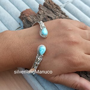 Op de afbeelding: Een zilveren manchetarmband met twee druppelvormige, lichtblauwe larimar stenen. De armband heeft een open ontwerp met sierlijke details rond de stenen. De tekst "silverRingManuco" is zichtbaar onderaan de afbeelding.