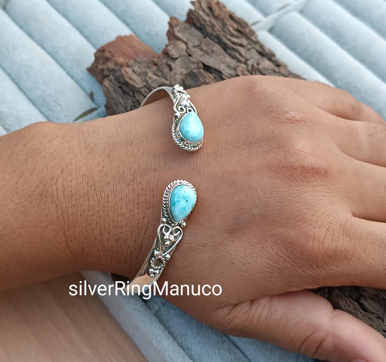 Op de afbeelding: Een zilveren manchetarmband met twee druppelvormige, lichtblauwe larimar stenen. De armband heeft een open ontwerp met sierlijke details rond de stenen. De tekst "silverRingManuco" is zichtbaar onderaan de afbeelding.