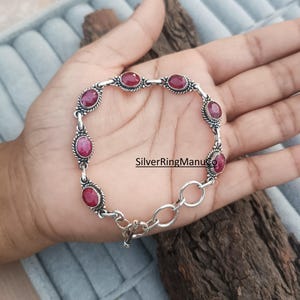 Könnte beinhalten: Ein silbernes Armband mit ovalen, tiefroten Edelsteinen. Das Armband hat ein dekoratives, gedrehtes Seildesign um jeden Stein und ist durch kleine Glieder verbunden. Der Verschluss ist ein einfacher Haken- und Ösenverschluss. Das Armband wird auf einer Hand präsentiert.