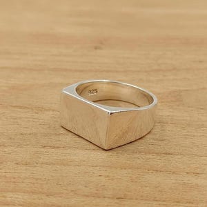 Könnte beinhalten: Ein polierter Siegelring aus Silber mit einer flachen, rechteckigen Vorderseite. Die Innenseite des Rings ist deutlich mit dem Stempel 925 versehen, was auf Sterlingsilber hinweist. Der Ring hat eine glatte, reflektierende metallische Oberfläche.