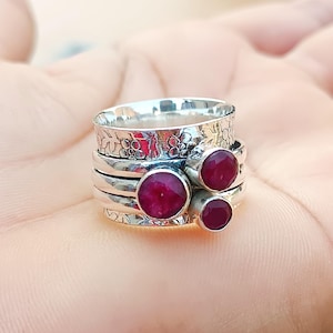 Könnte beinhalten: Ein silberner Drehring mit drei roten Edelsteinen. Der Ring ist mit einem Blumenmuster verziert.