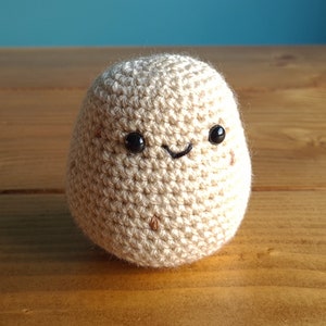 May include: A crocheted potato toy with a smiling face. The potato is beige and has black eyes and a mouth.